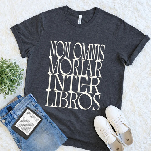 A dark heather gray graphic t-shirt with the phrase 'NON OMNIS MORIAR INTER LIBROS' printed in large cream serif letters. The t-shirt is styled in a flat lay on a white faux fur background, accompanied by blue jeans, white sneakers, and a small green plant.