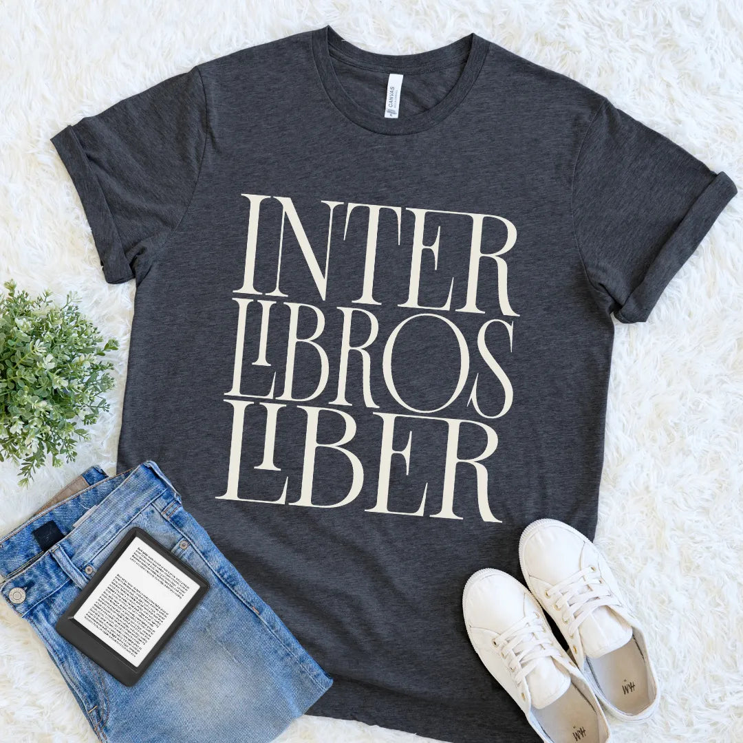 A dark charcoal gray bookish graphic t-shirt with the text 'INTER LIBROS LIBER' in a cream serif font, styled in a flat lay with blue jeans, white sneakers, and a small green plant on a white faux fur background.