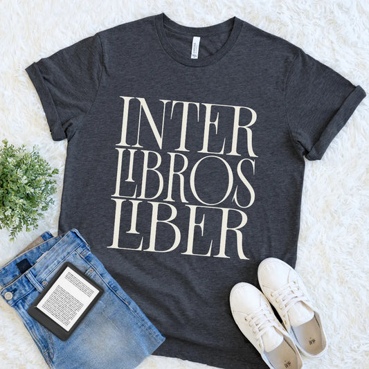 A dark charcoal gray bookish graphic t-shirt with the text 'INTER LIBROS LIBER' in a cream serif font, styled in a flat lay with blue jeans, white sneakers, and a small green plant on a white faux fur background.