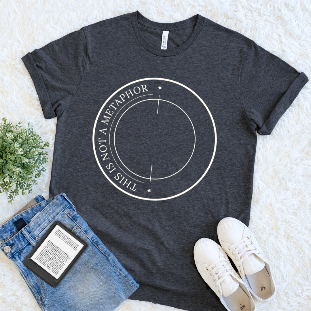 A dark heather gray graphic t-shirt for literature students with a white circular design reading 'THIS IS NOT A METAPHOR' is styled in a flat lay on a white faux fur background, accompanied by blue jeans, white sneakers, and a small green plant