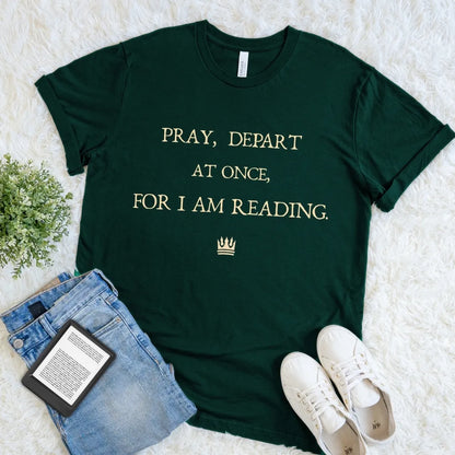 A dark forest green graphic t-shirt with gold text reading 'PRAY, DEPART AT ONCE, FOR I AM READING.' and a small crown icon, styled in a flat lay on a white faux fur background with jeans, white sneakers, a book, and a small plant.