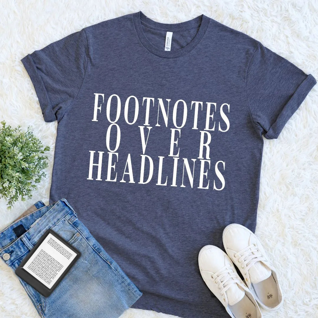 A heather navy blue graphic t-shirt with the text 'FOOTNOTES OVER HEADLINES' in white serif letters, styled in a flat lay with blue jeans, white sneakers, a book, and a small green plant on a white shag rug.