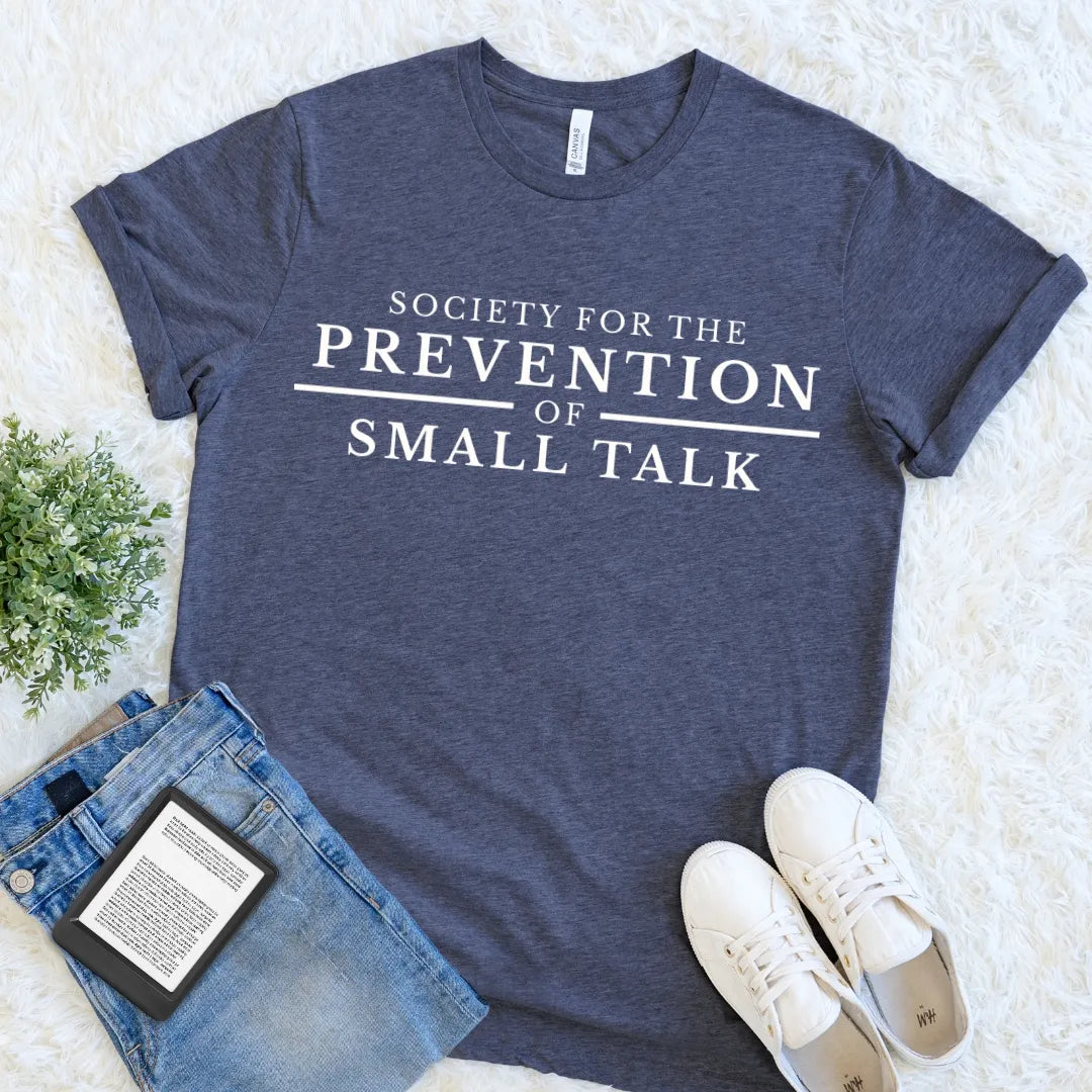 A heather navy blue graphic t-shirt with white text reading 'SOCIETY FOR THE PREVENTION OF SMALL TALK', styled in a flat lay on a white faux fur rug with blue jeans, white sneakers, and a small green plant.