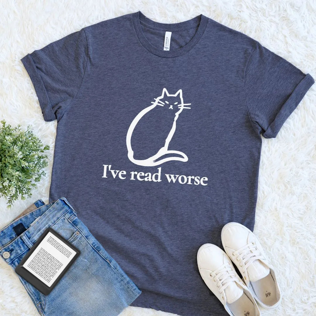 A heather navy blue bookish graphic t-shirt with a white line art cat and the text 'I've read worse' printed on the front. The t-shirt is styled in a flat lay on a white faux fur background, accompanied by blue jeans, a black Kindle e-reader, and white sneakers.