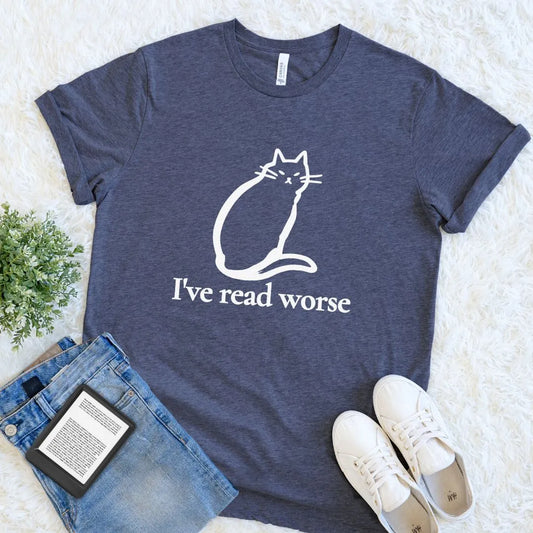 A heather navy blue bookish graphic t-shirt with a white line art cat and the text 'I've read worse' printed on the front. The t-shirt is styled in a flat lay on a white faux fur background, accompanied by blue jeans, a black Kindle e-reader, and white sneakers.