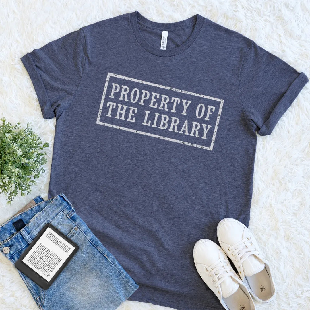 A heather navy blue reader t-shirt with a distressed white graphic reading 'PROPERTY OF THE LIBRARY' laid flat on a white faux fur background, styled with blue jeans, a book, and white sneakers.