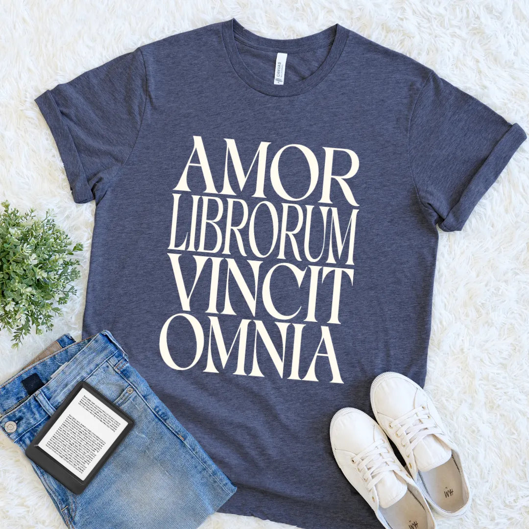 A heather navy blue vintage inspired reader shirt with the phrase 'AMOR LIBRORUM VINCIT OMNIA' printed in large white serif letters, styled in a flat lay with blue jeans, white sneakers, a book, and a small green plant on a white faux fur background.