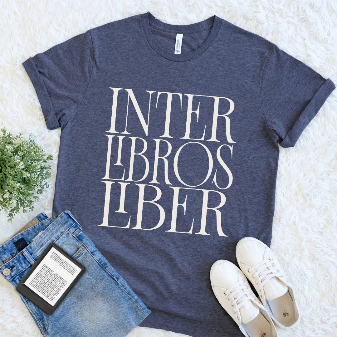 A heather navy blue bookish graphic t-shirt with the words 'INTER LIBROS LIBER' printed in large white serif letters, styled in a flat lay on a white faux fur background with blue jeans, a book, and white sneakers.