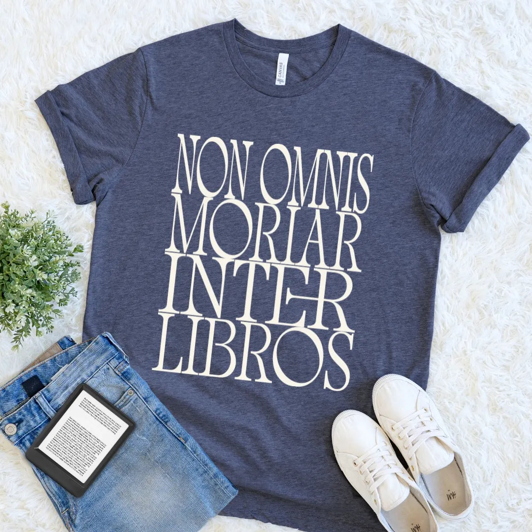 Dark academia reader navy blue heather  t-shirt with the phrase 'NON OMNIS MORIAR INTER LIBROS' printed in large white serif letters. The t-shirt is styled in a flat-lay arrangement on a white faux fur background, accompanied by blue jeans, a black Kindle e-reader, and white sneakers.