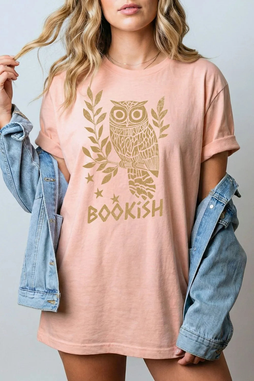 Person wearing a heather peach reader  t-shirt with an owl design and 'bookish' text, holding a denim jacket for relaxed reading outfit