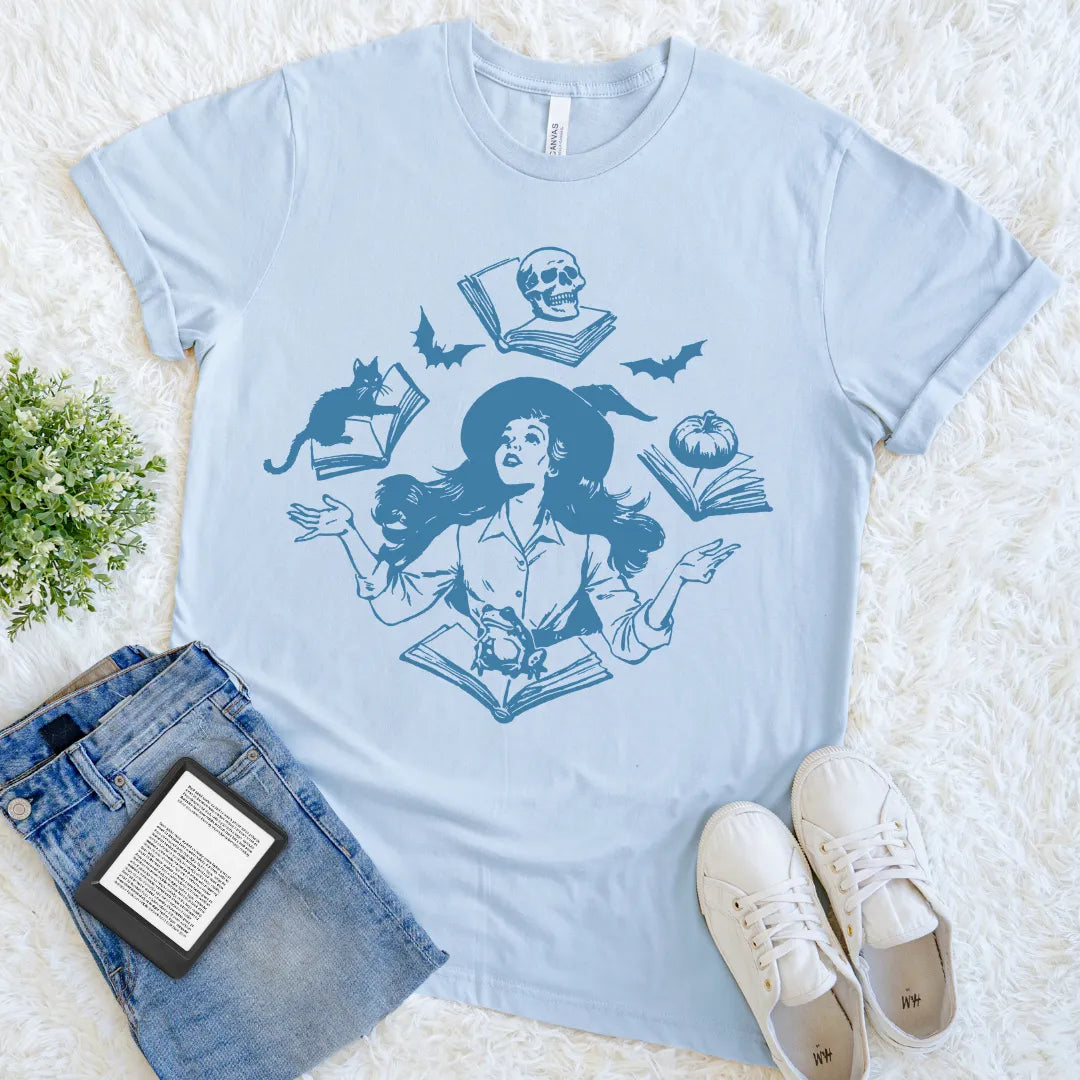 Light blue halloween t-shirt with a graphic design of a witch, cat, and books on a white surface.