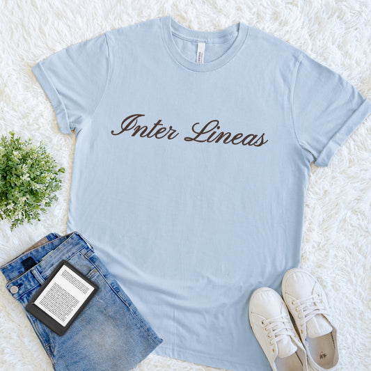 A light blue crewneck t-shirt with the text "Inter Lineas" — Latin for between the lines printed in dark brown serif letters across the chest. The t-shirt is styled in a flat lay on a white faux fur background, accompanied by blue jeans, white sneakers, and a small potted green plant.