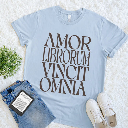 A light blue book lover graphic t-shirt with the text 'AMOR LIBORUM VINCIT OMNIA' Latin typography printed in dark brown serif letters. The t-shirt is styled in a flat lay on a white faux fur background, accompanied by blue jeans, white sneakers, and a small green plant.