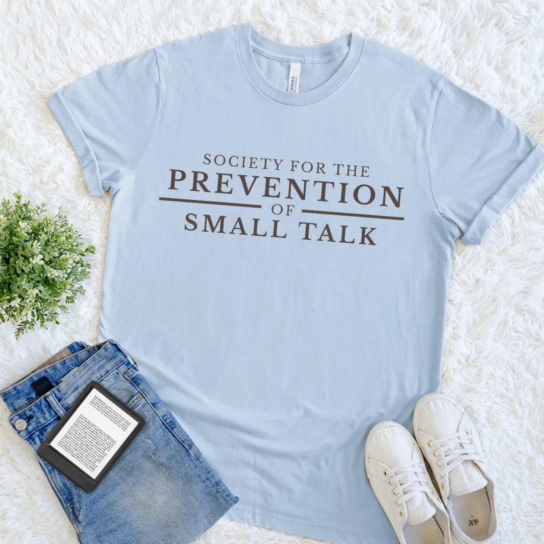 A light blue graphic t-shirt with a minimalist academic quote design for readers   'SOCIETY FOR THE PREVENTION OF SMALL TALK' in dark brown lettering, styled in a flat lay with blue jeans, white sneakers, and a small potted plant on a white faux fur background.