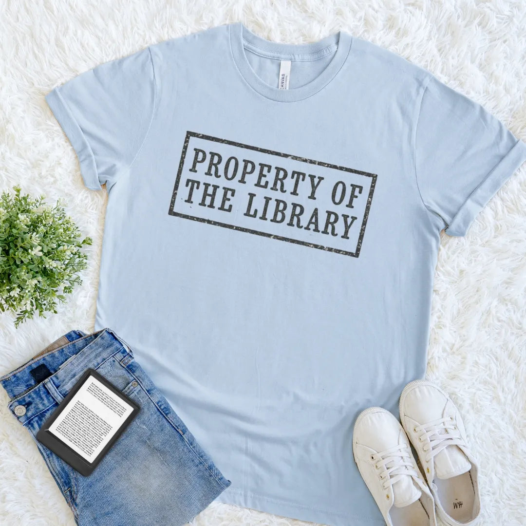 A light blue crewneck bookish t-shirt featuring a distressed rectangular library ownership stamp design reading 'PROPERTY OF THE LIBRARY' in a rectangular border. The shirt is styled in a flat lay on a white faux fur rug alongside blue jeans, white sneakers, a book, and a small green plant.