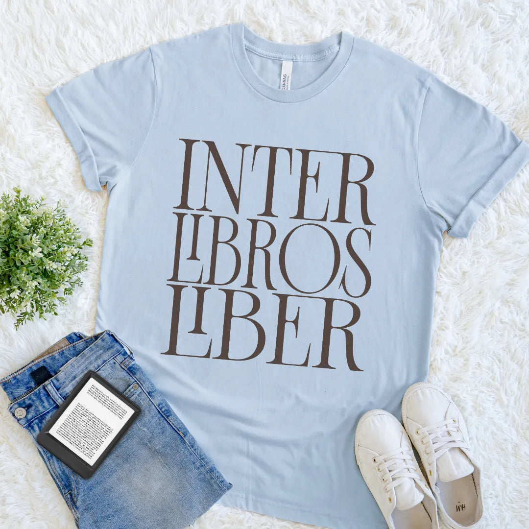 A light blue graphic t-shirt for book lovers with the text 'INTER LIBROS LIBER' printed in dark brown serif letters, styled in a flat lay on a white faux fur rug with blue jeans, white sneakers, and a small potted plant.