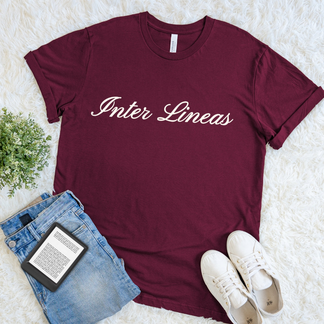 A maroon short-sleeve t-shirt with 'Inter Lines' printed in white script across the chest, styled in a flat lay with blue jeans, white sneakers, and a small green plant on a white faux-fur background.