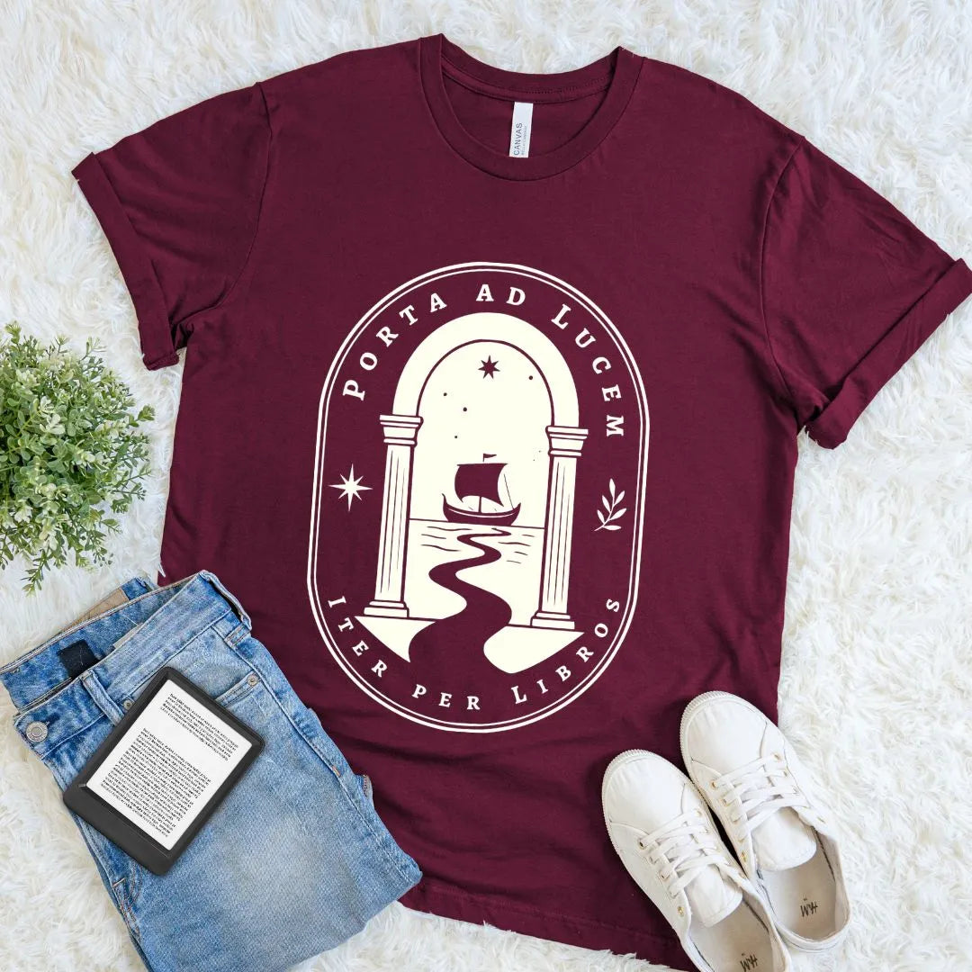 Maroon Porta ad Lucem book lover T-shirt with ivory vintage archway, sailing ship, stars, and literary Latin-inspired graphic, styled flat with jeans and a book.