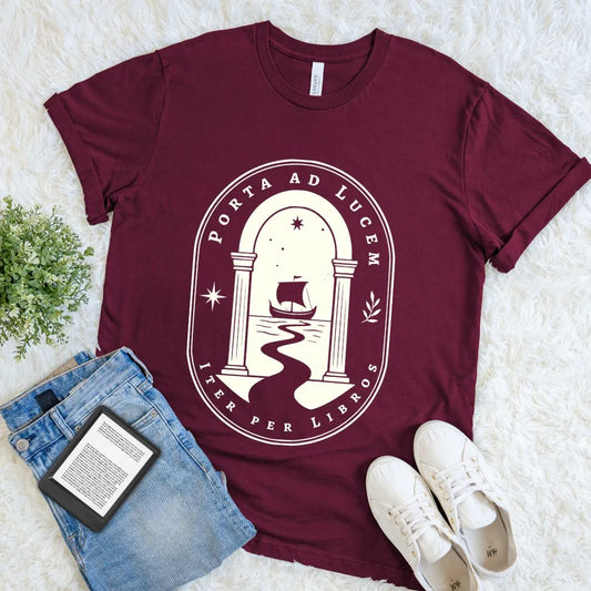 Bookish T shirt with ancient archway, sailing ship, and stars in vintage style design on maroon red cotton, styled with jeans and white sneakers on white surface