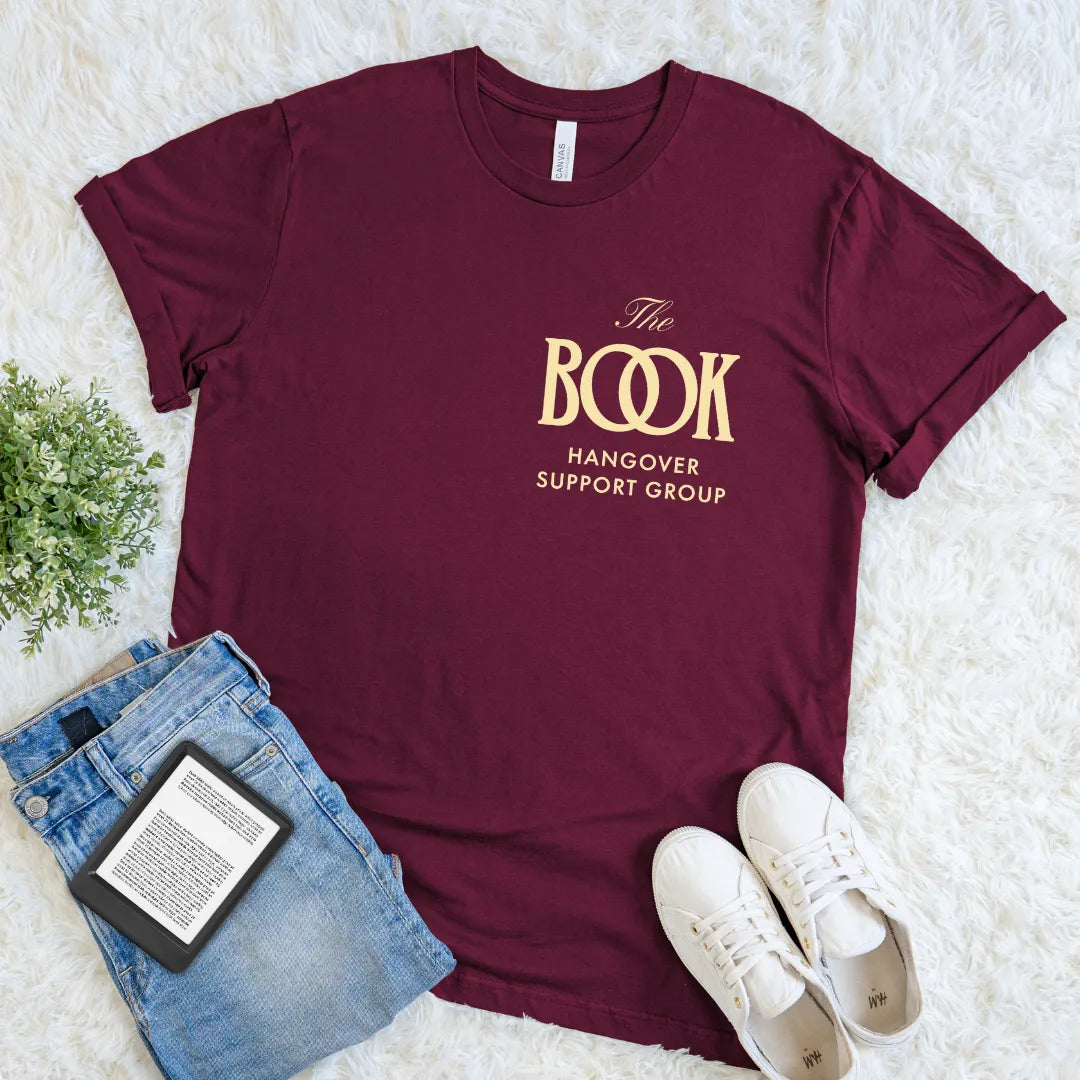 Flat lay of maroon red soft cotton t-shirt   featuring fashion logo with text “The Book Hangover Support Group” in elegant lettering in gold