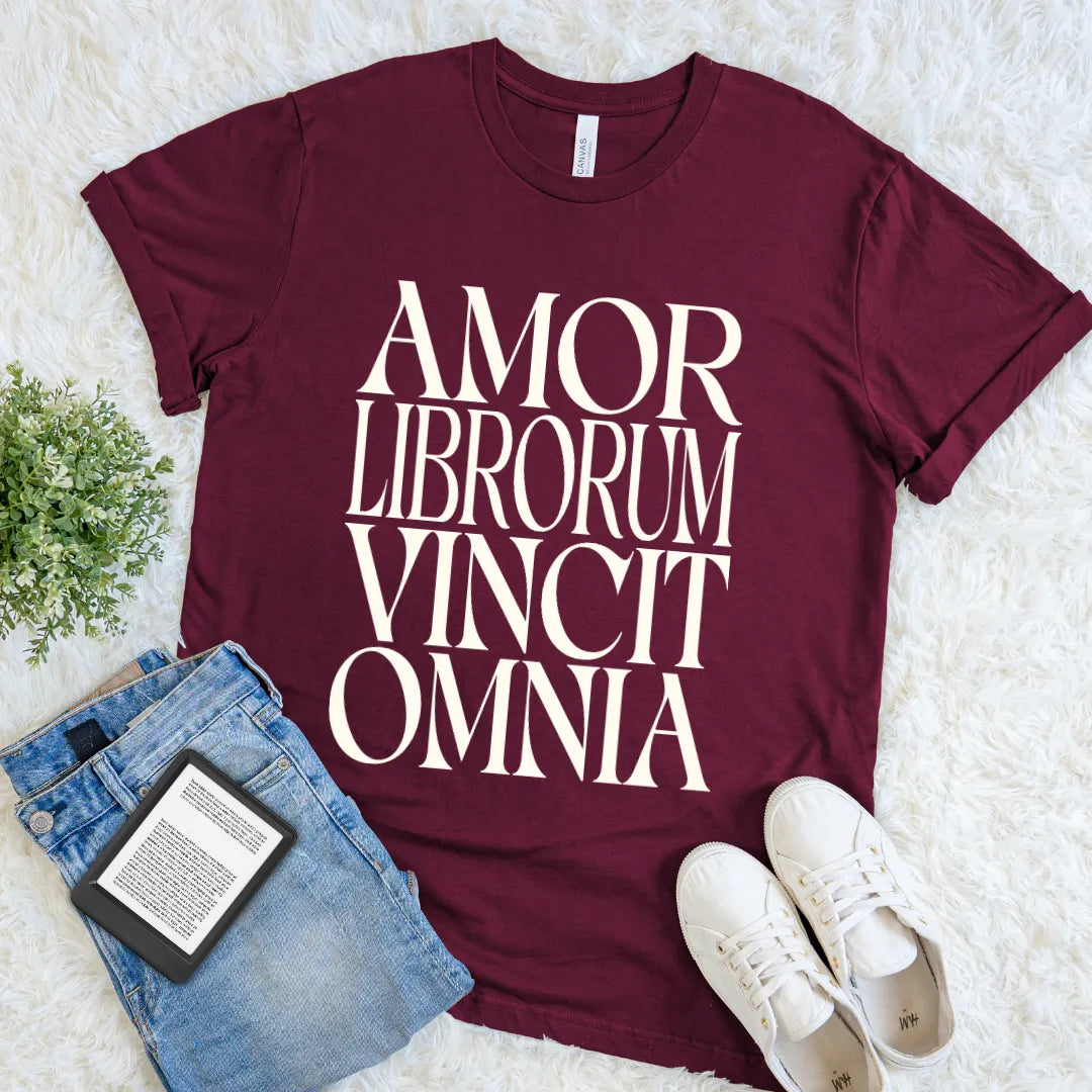 A maroon short-sleeve graphic T-shirt laid flat on a white faux-fur surface, printed with large white serif text reading 'AMOR LIBRORUM VINCIT OMNIA.' meaning love of books conquers all in elegant typography. Folded blue jeans, white sneakers, and a small potted plant surround it as styling props.