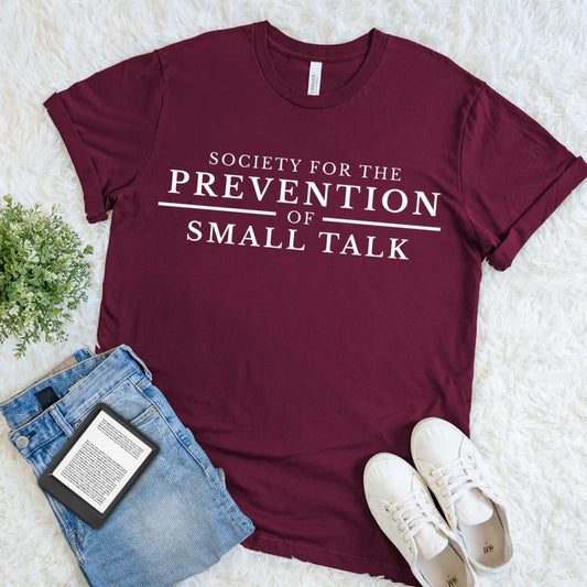 A maroon short-sleeve graphic T-shirt for introverts laid flat on a white faux-fur surface, styled with blue jeans, white sneakers, and a small green plant. The shirt has large white text reading 'SOCIETY FOR THE PREVENTION OF SMALL TALK.