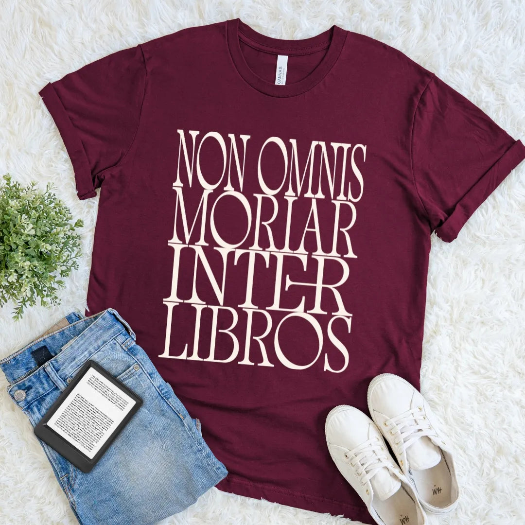 A maroon short-sleeve graphic T-shirt laid flat on a white faux-fur surface, styled with blue jeans, white sneakers, and a small green plant. The shirt has large Bookish Latin quote with a classic literary design for devoted readers and bibliophiles reading 'NON OMNIS MORIAR INTER LIBROS'.