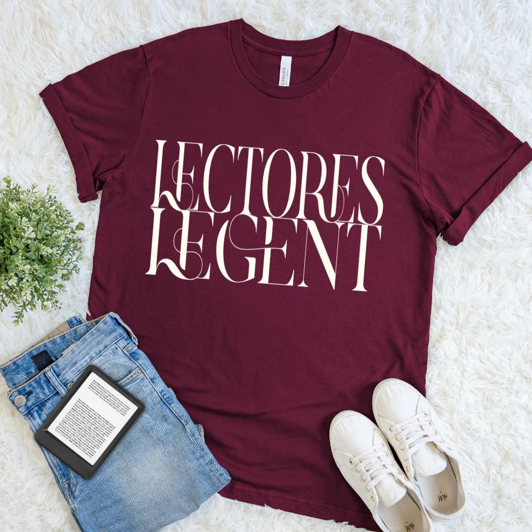A maroon short-sleeve graphic T-shirt for dark academia fans with  the words 'LECTORES LEGENT' means readers gonna read printed in large white serif letters across the front. The shirt is styled in a flat lay on a white faux-fur surface with blue jeans, white sneakers, a small green plant, and a book.