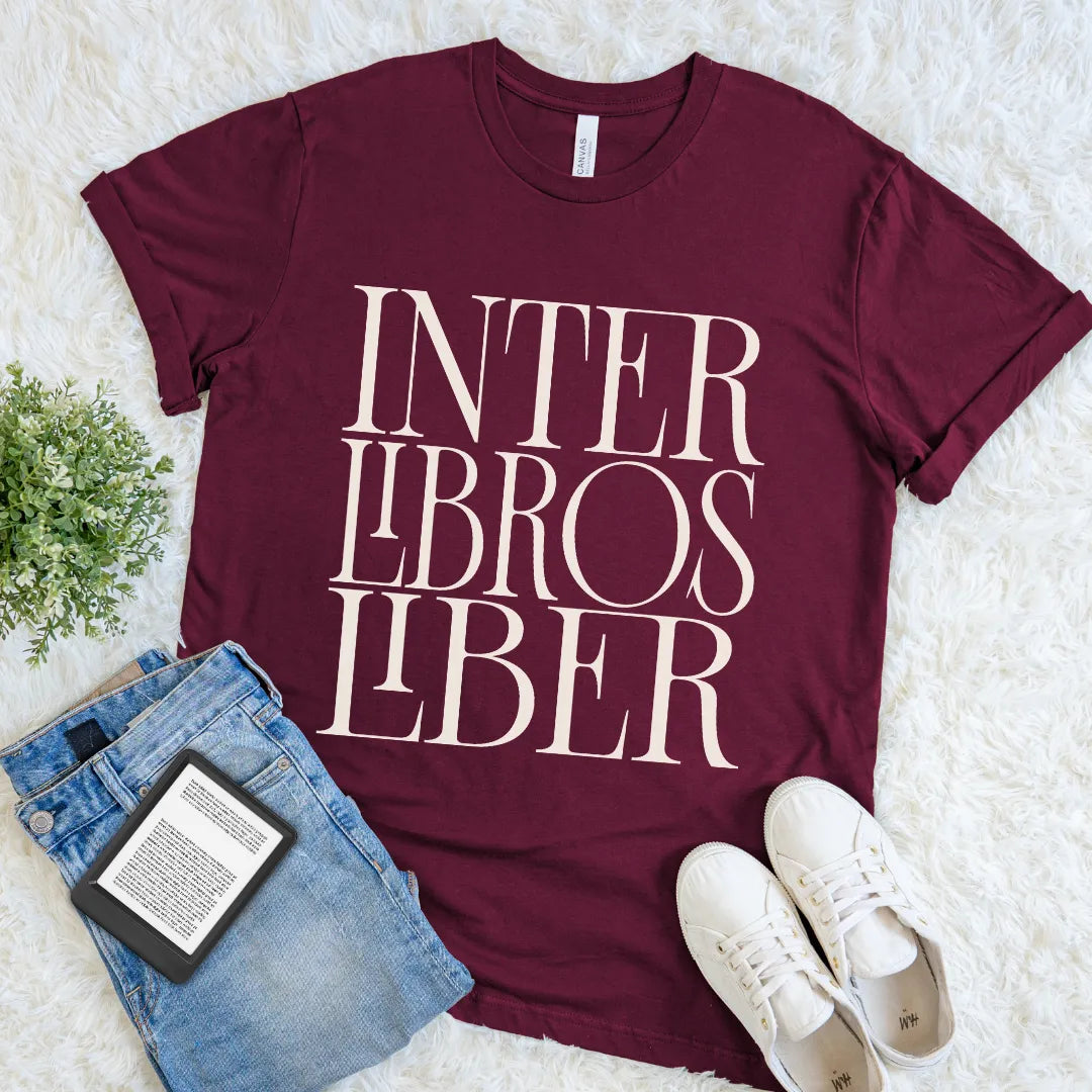 A maroon short-sleeve graphic T-shirt for book lovers laid flat on a white faux-fur surface, styled with blue jeans, a book, and white sneakers. The shirt has large cream text reading 'INTER LIBROS LIBER' across the front.
