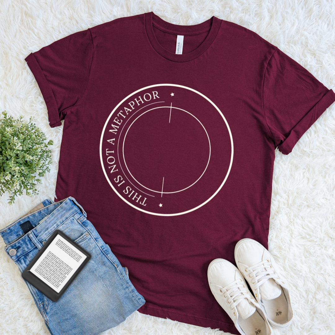 A maroon red bookish reader tee with witty metaphor quote and academic-inspired circular line art laid flat on a white faux-fur surface, styled with blue jeans, white sneakers, and a small green plant. The shirt has a white circular line graphic with the text 'THIS IS NOT A METAPHOR' around the center.