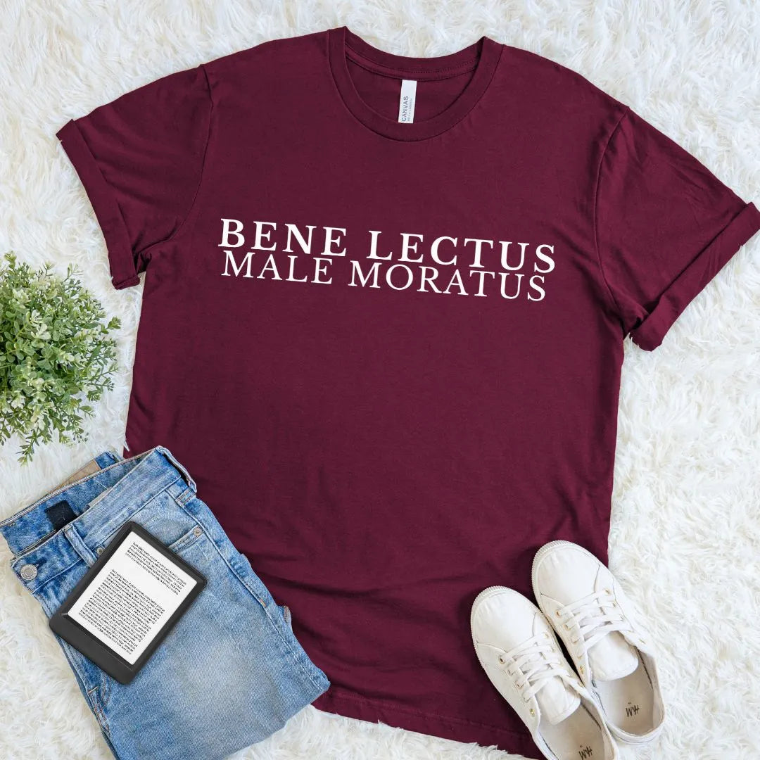 A maroon short-sleeve graphic T-shirt with rolled sleeves and the text 'BENE LECTUS MALE MORATUS' in minimalist vintage typography printed in white across the chest, styled in a flat lay with blue jeans, white sneakers, a book, and a small green plant on a white textured background.
