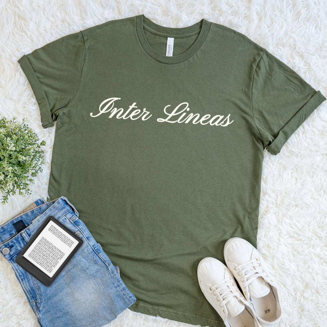 An olive green short-sleeve t-shirt featuring the Latin phrase Inter Lineas in elegant brown script lettering  printed in cream across the chest, styled in a flat lay on a white faux fur background with blue jeans, white sneakers, and a small potted plant.