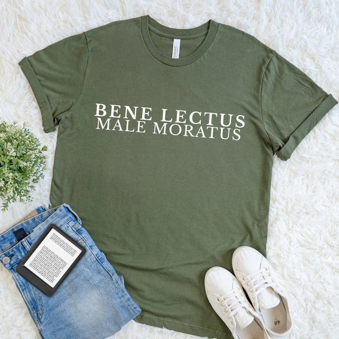 An olive green graphic t-shirt with the text 'Bene Lectus Male Moratus Latin quote in cream serif typography, styled in a flat lay on a white faux fur rug with blue jeans, a book, and white sneakers.