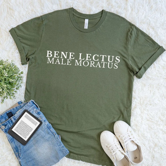 An olive green graphic t-shirt with the text 'Bene Lectus Male Moratus Latin quote in cream serif typography, styled in a flat lay on a white faux fur rug with blue jeans, a book, and white sneakers.