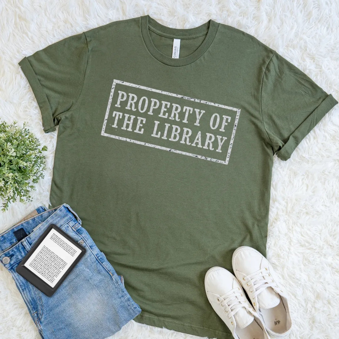 An olive green short-sleeve t-shirt with a distressed white graphic reading 'PROPERTY OF THE LIBRARY' in a rectangular stamp design. The shirt is styled in a flat lay on a white faux fur rug alongside blue jeans, a book, and white sneakers.