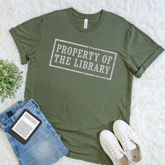 An olive green short-sleeve t-shirt with a distressed white graphic reading 'PROPERTY OF THE LIBRARY' in a rectangular stamp design. The shirt is styled in a flat lay on a white faux fur rug alongside blue jeans, a book, and white sneakers.
