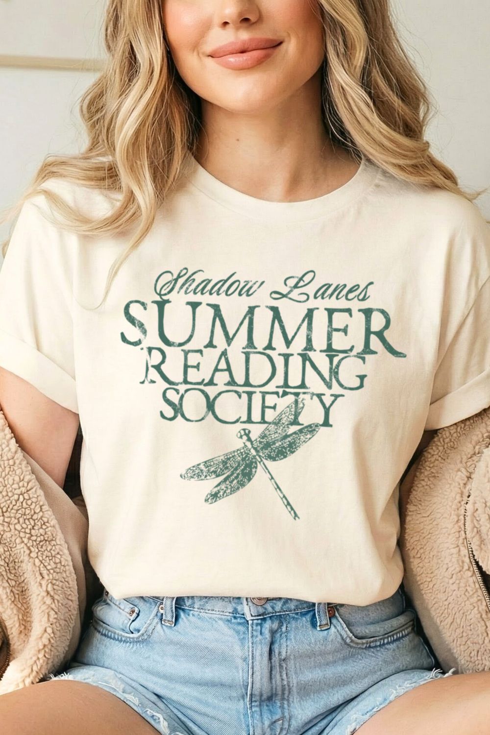woman wearing a natural beige shirt with  'Shadow Lanes Summer Reading Society' text with a dragonfly graphic styled with jeans for cozy bookish outfit.