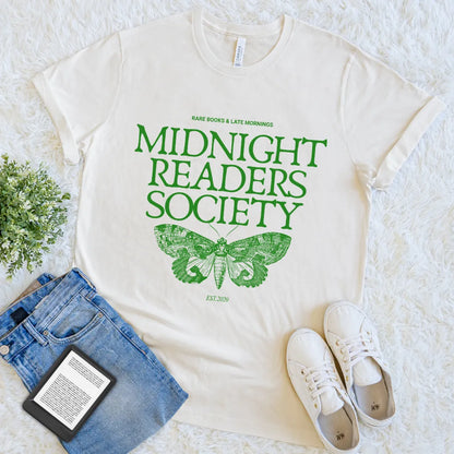Flat lay of the soft cotton cream t-shirt with green moth design and "midnight readers society" quote, styled with denim  and e-reader for bookish look
