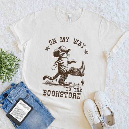 Soft cotton cream tee featuring Western-style design with walking cat and desert elements and "on my way to the bookstore" text