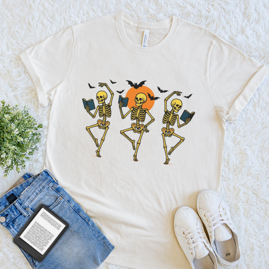 A cream-colored short-sleeve bookish halloween t-shirt with a graphic of three dancing skeletons holding books in front of an orange sun and black bats. The shirt is styled in a flat lay on a white textured rug with blue jeans, white sneakers, and a small green plant.