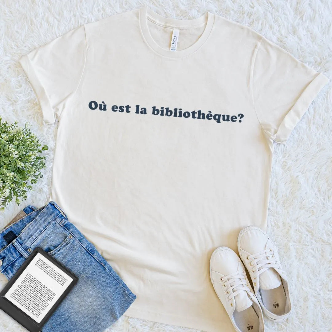 A natural beige  short-sleeve graphic T-shirt laid flat on a white textured rug, with the French text 'Où Est La Librairie?' printed across the chest in dark navy. Styled flat-lay props include blue jeans, white sneakers, a book, and a small green plant.
