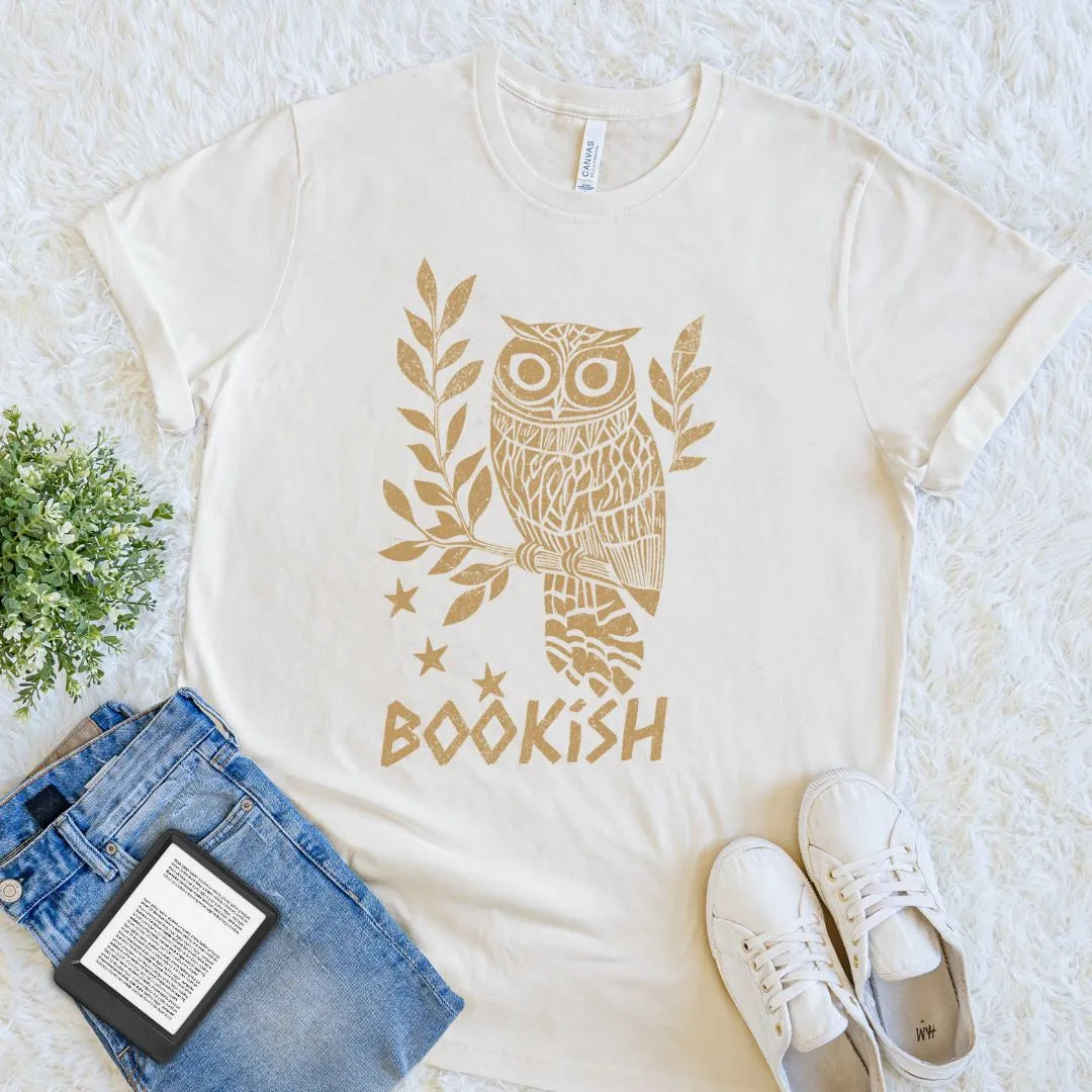 Natural beige t-shirt with owl design and 'bookish' text, placed on a textured surface with jeans, sneakers, and an e-reader.
