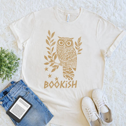 Natural beige t-shirt with owl design and 'bookish' text, placed on a textured surface with jeans, sneakers, and an e-reader.