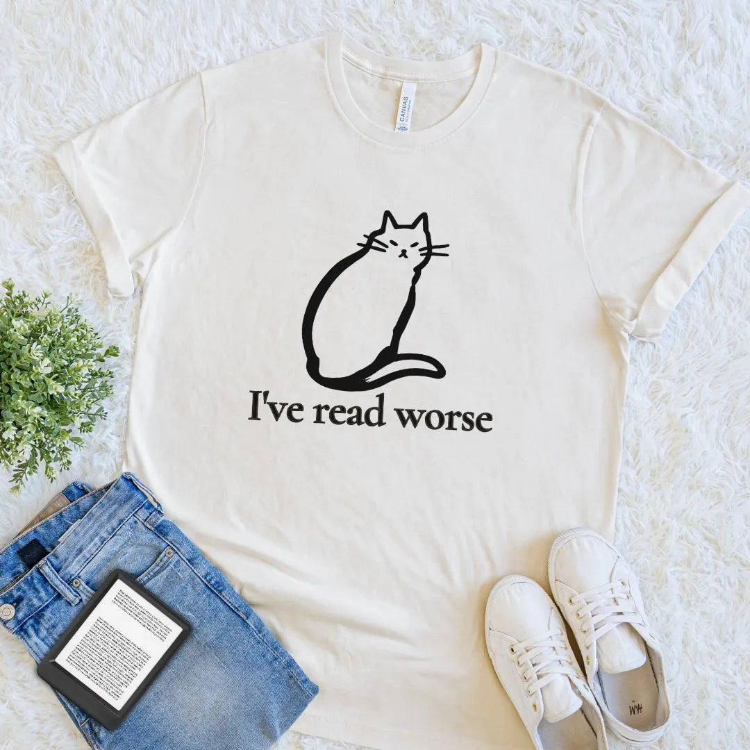 An off-white cream bookish graphic t-shirt featuring a modern minimalist design with black line art cat and the humorous quote 'I've read worse', styled in a flat lay with blue jeans, white sneakers, a book, and a small green plant on a white textured rug.