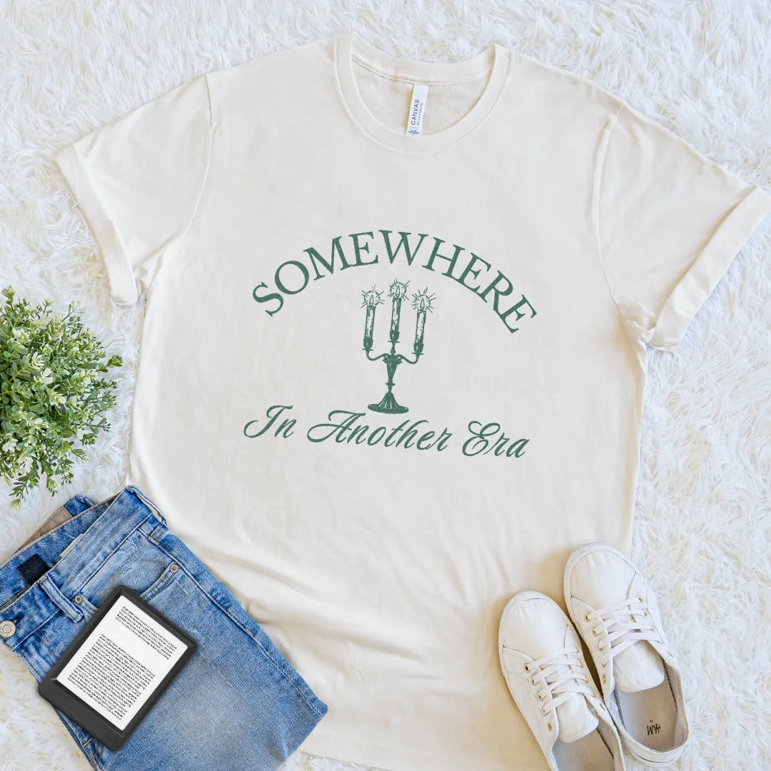 Cream bookish shirt with vintage candle illustration and elegant serif text  'Somewhere In Another Era' in muted green on a textured surface with jeans and sneakers.