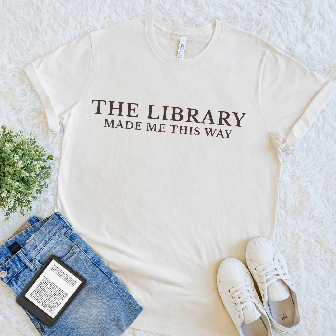 An off-white graphic t-shirt with The Library Made Me This Way reader identity statement in dark brown lettering, styled in a flat lay with blue jeans, white sneakers, a book, and a small green plant on a white textured rug.