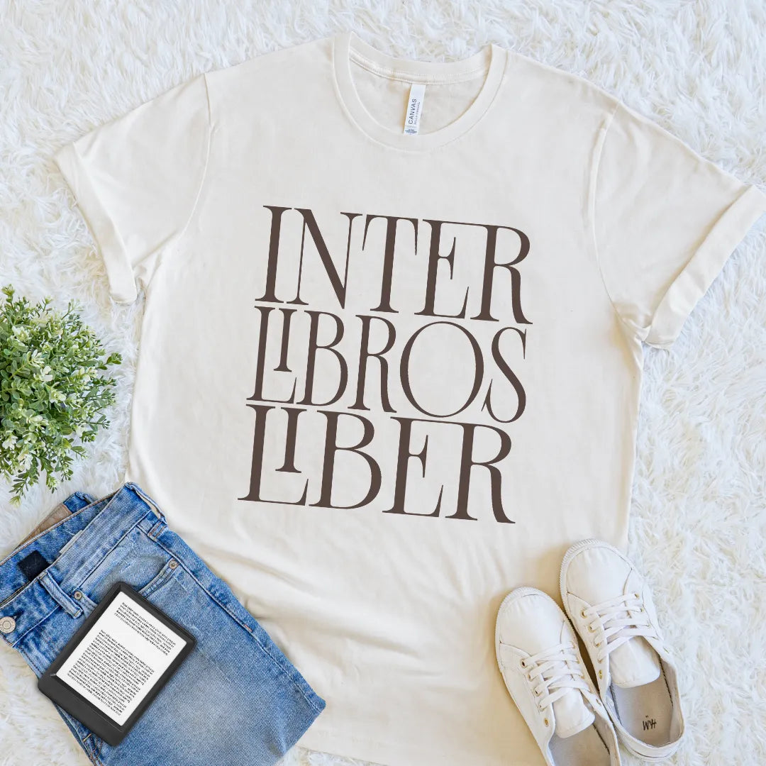 An off-white crewneck reader t-shirt with the text 'INTER LIBROS LIBER' printed in dark brown serif letters, styled in a flat lay with blue jeans, white sneakers, a book, and a small green plant on a white textured rug.
