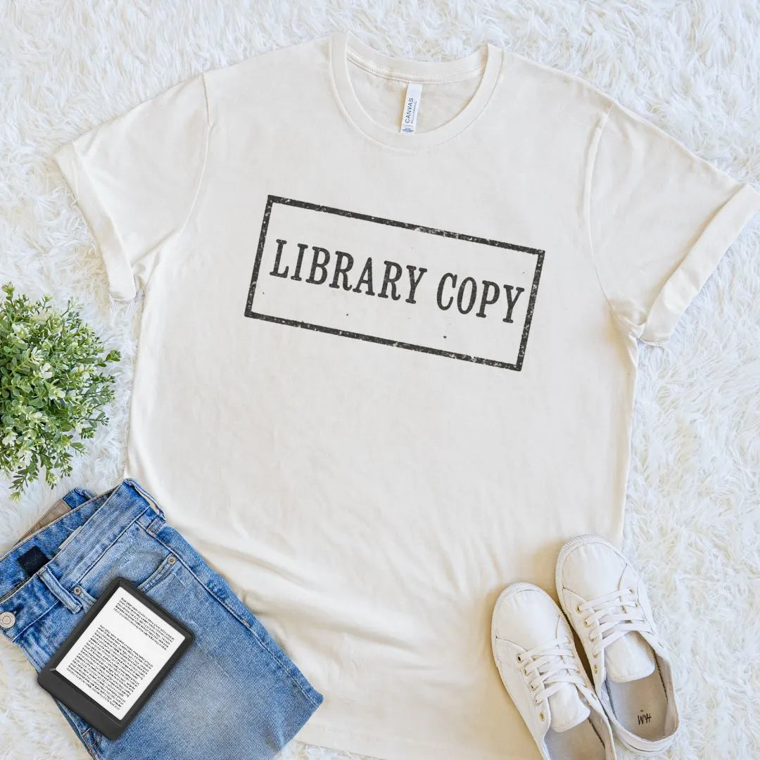 An natural beige bookish crewneck t-shirt with a black distressed 'LIBRARY COPY' stamp graphic, styled in a flat lay with blue jeans, white sneakers, a book, and a small green plant on a white shag rug.