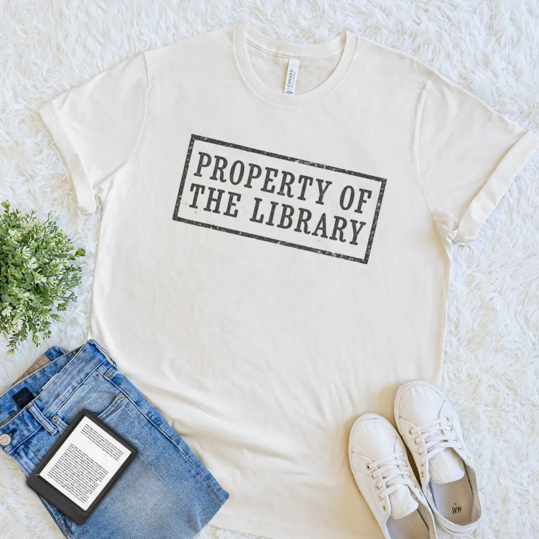 A natural beige crewneck t-shirt with a distressed black stamp-style graphic reading 'PROPERTY OF THE LIBRARY', styled in a flat lay on a white faux fur rug with blue jeans, white sneakers, a book, and a small green plant.