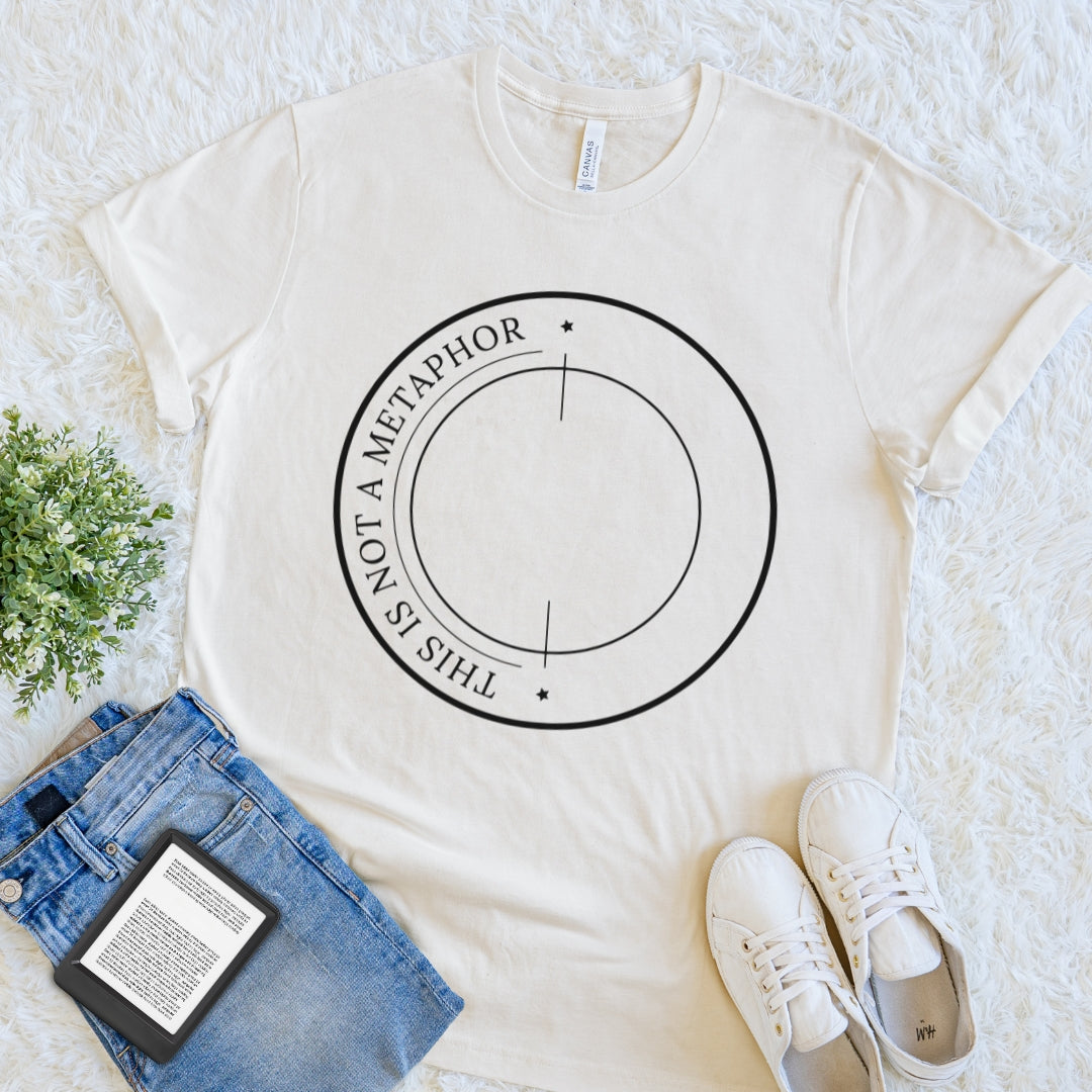 An off-white crewneck t-shirt  for readers with a black circular graphic reading 'THIS IS NOT A METAPHOR' is styled in a flat lay on a white textured rug, accompanied by blue jeans, white sneakers, a book, and a small green plant.